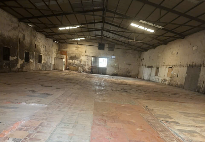 kho ccn an khánh Interior of a large empty warehouse with peeling walls, exposed metal roof trusses, and a dusty tiled floor.